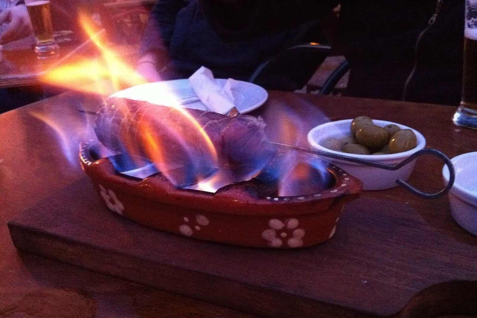Eine geräucherte Wurst, die in einer rot gemusterten Tonschale auf einem Holzbrett flammt, daneben Oliven und ein Glas
