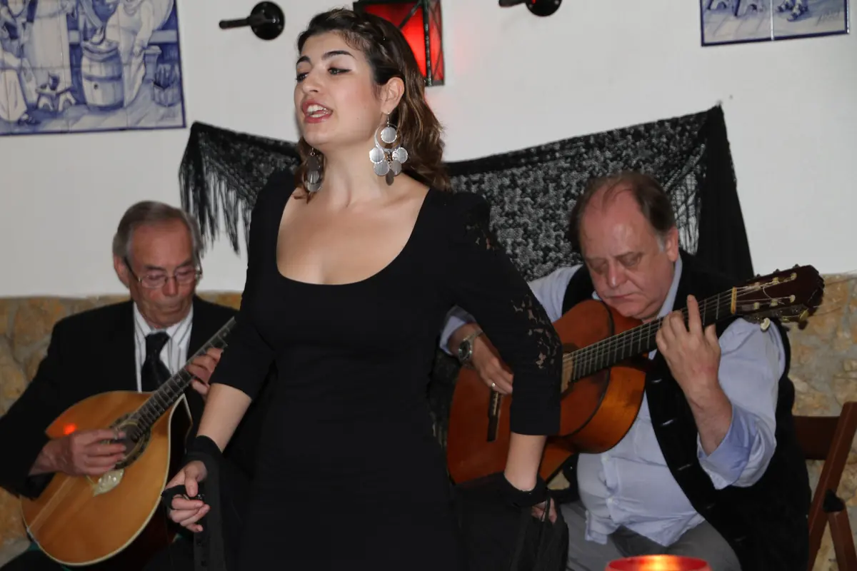 Fado zpěvačka v černých šatech mezi dvěma kytaristy, jeden drží hruškovitou portugalskou kytaru a druhý šestistrunnou kytaru, v malém lisabonském fado domě s modrými kachlíky a červenou lucernou na zdi