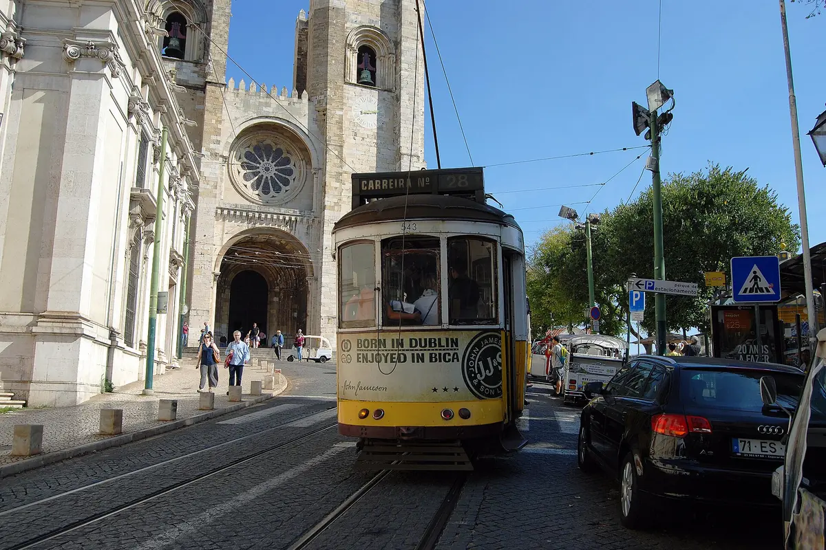 Žlutá tramvaj projíždí pod dvojicí věží lisabonské katedrály Sé na slunné ulici