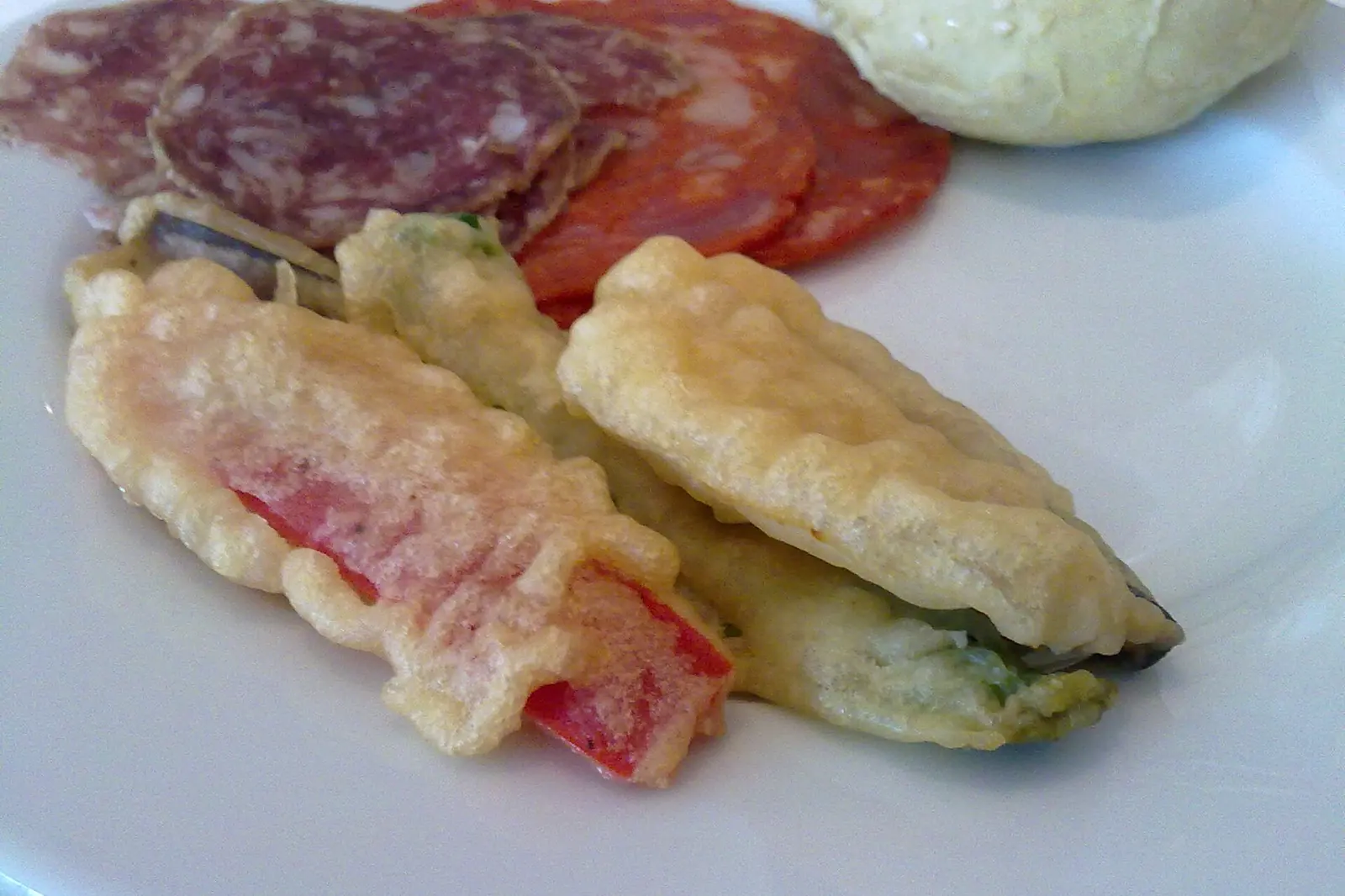 Ein weißer Teller mit frittiertem grünen Gemüse im Teigmantel neben Scheiben von luftgetrockneter Wurst und einem Brötchen