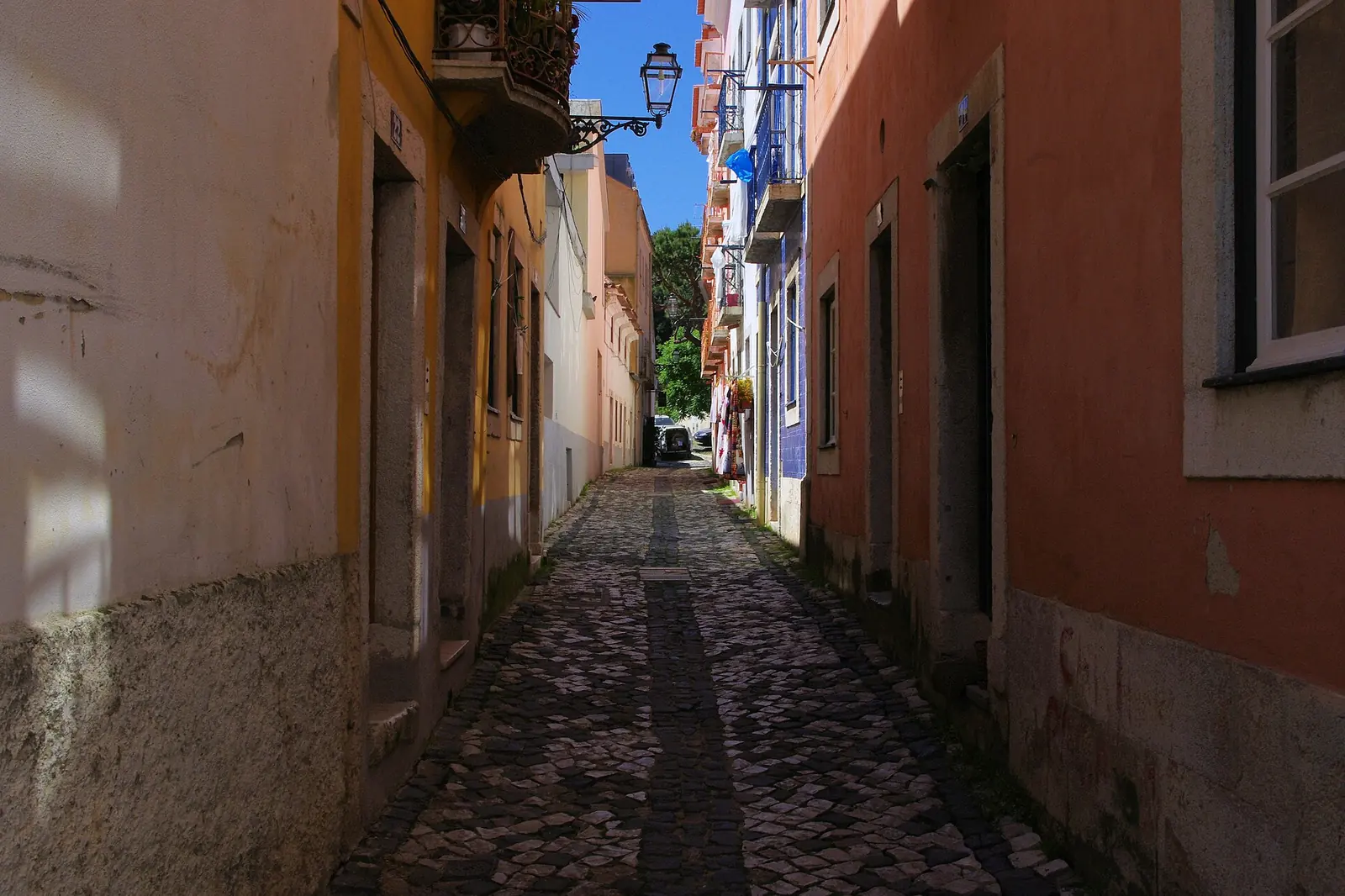 Eine enge Gasse im Alfama-Viertel von Lissabon, wo ein verwittertes Holzhaus auf einer Steinmauer über einem kleinen Platz mit Tauben aufragt