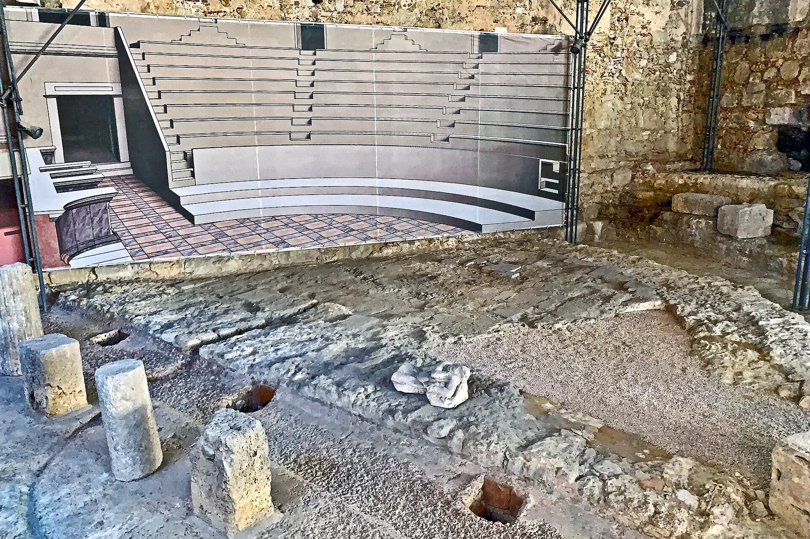 Freigelegte Steinstufen und Mauern des römischen Theaters von Lissabon unter einem schützenden Stahldach, im Hintergrund ein Modell der Bühne